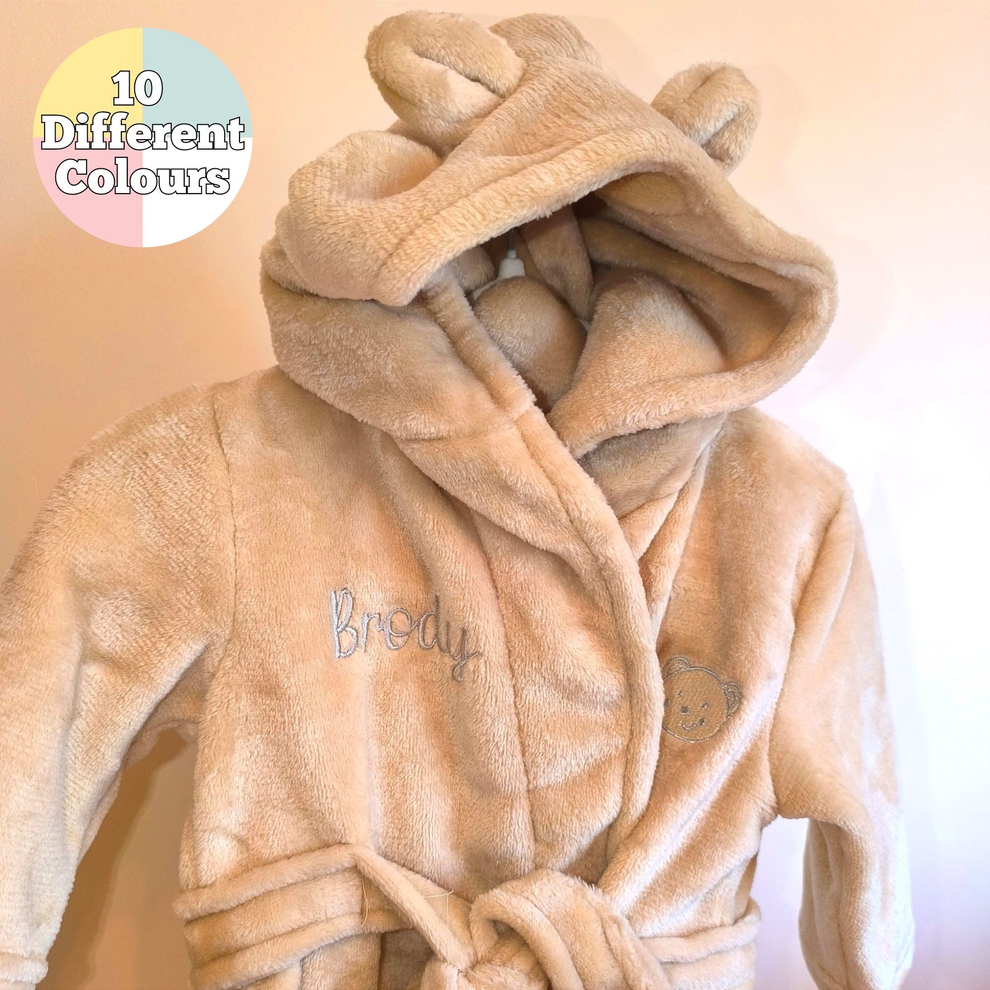 A personalised baby dressing gown in a beige color with bear ears on the hood, embroidered name on the chest, and a grey hoodie with a bear face on the left chest.