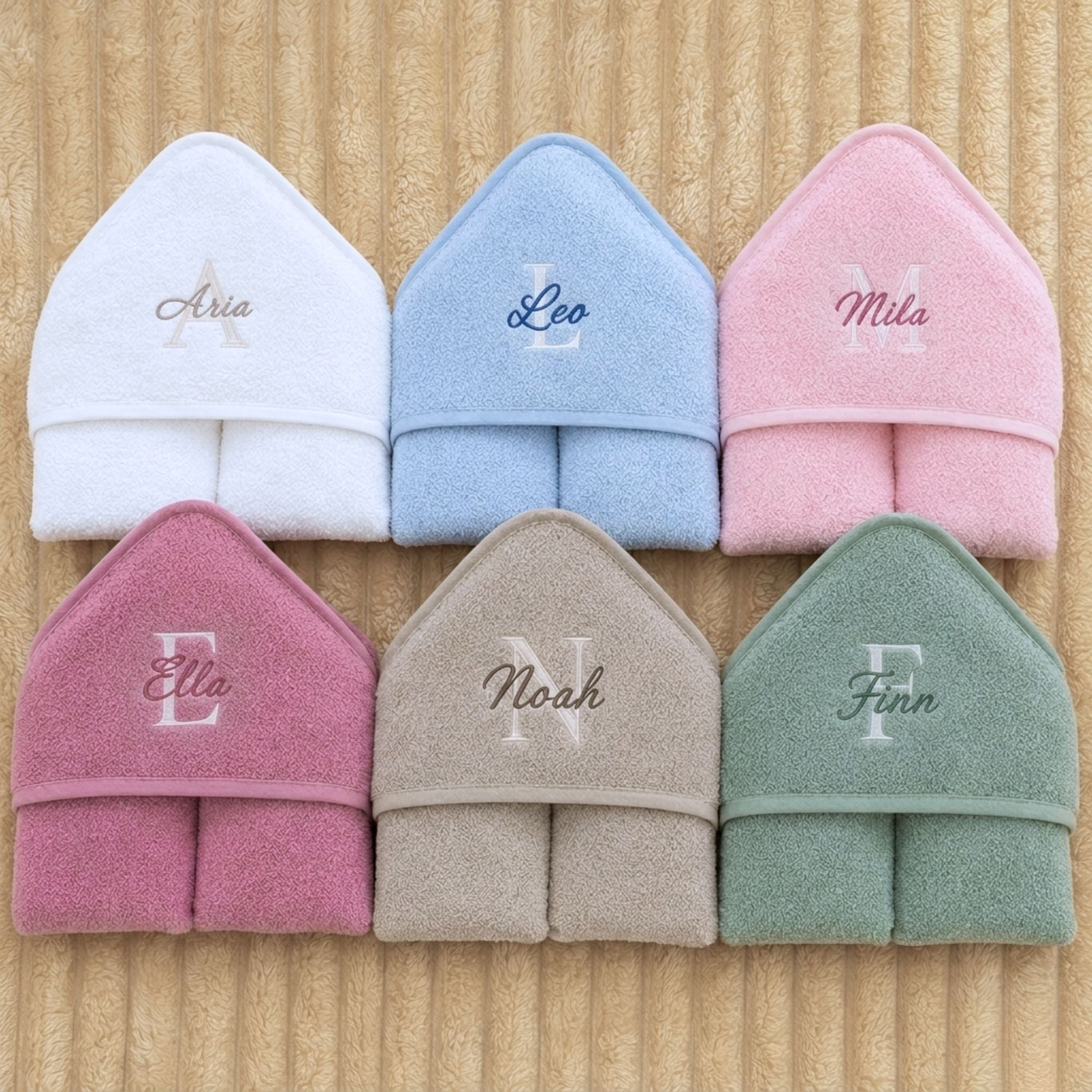 Set of six folded towels in white, blue, pink, red, beige, and green with embroidered names on a wooden surface.