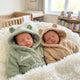 Two babies in bear-ear dressing gowns with a personalised name embroidered on the chest, lying on a fluffy blanket in a nursery.