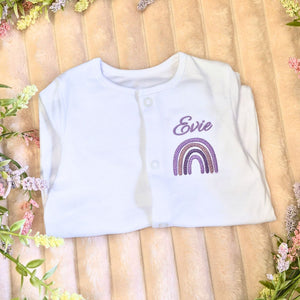 personalised baby name sleepsuit in white with an embroidered lilac rainbow design and a baby name above.